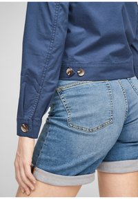 Gros plan d'une personne portant un short en jean bleu retroussé et une veste bleue à manches longues avec des boutons aux poignets et à la taille.