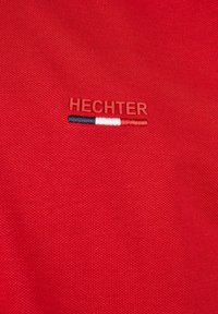 Roter Stoff mit strukturierter Webart, ausgestattet mit einem gestickten "HECHTER"-Logo in passendem Rot und zwei horizontalen Streifen in Schwarz und Weiß.