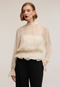 Blusa color beige chiaro, trasparente con design plissettato, collo alto e orlo con volant. Maniche lunghe con polsini arricciati, indossata sopra pantaloni neri.