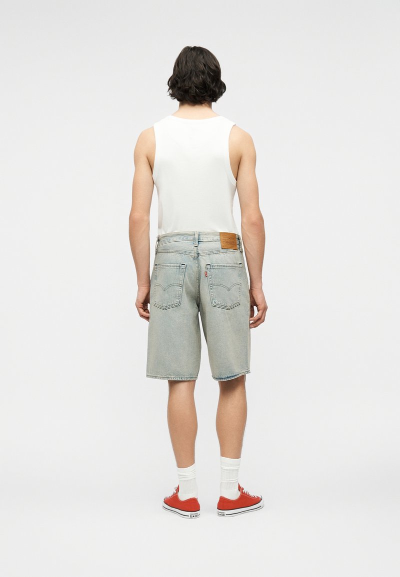 Mann trägt weißes ärmelloses Hemd, hellblaue Jeansshorts, weiße Socken und rote Turnschuhe, steht mit dem Rücken zur Kamera vor einfachem Hintergrund.