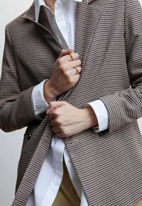 Blazer de patrón pata de gallo en tonos marrón y beige, tela texturizada, cierre con un solo botón, llevado sobre una camisa blanca, manos agarrando la chaqueta.