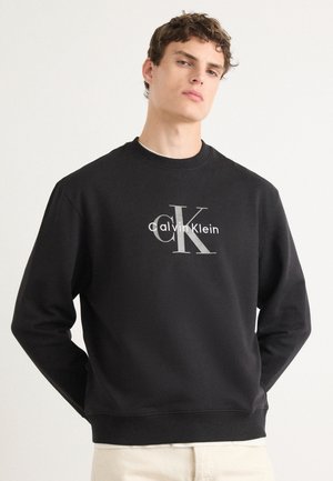 Jeune homme portant un sweat-shirt noir Calvin Klein avec un grand logo "cK" et un texte blanc, debout devant un fond clair uni.