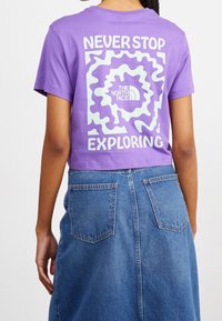 Purple cropped t-shirt featuring white text "NEVER STOP EXPLORING" and a swirl design, paired with a high-waisted blue denim skirt.