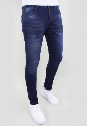 Person iført mørkeblå skinny jeans og hvide sneakers, stående mod en ensfarvet lys baggrund.