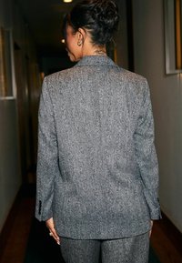 Blazer grigio strutturato con un motivo a spina di pesce. La parte posteriore presenta un taglio classico e bottoni semplici, completato da un paio di pantaloni abbinati.