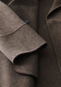 Bruine, geaccentueerde wollen jas met een zwarte knop en pipingdetail op een gevouwen voorkant.