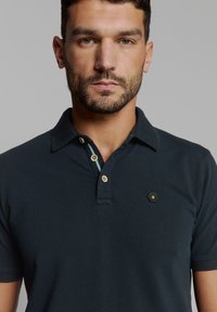 Donker marineblauwe poloshirt met een tweeknoopsluiting, goudkleurige knopen en een zeshoekig embleem op de linkerkant van de borst. Gemaakt van katoen met een gladde textuur.