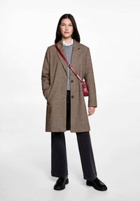 Svetlo rjav in krem karirast plašč z izrezanim ovratnikom, zapiranjem na en gumb in stranskimi žepi, oblečen čez sivo pulover in črne hlače.