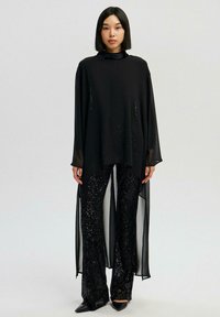 Sort flydende tunika med lange ærmer og høj hals, kombineret med sorte flæsebukser med palietter og spidsede hæle.