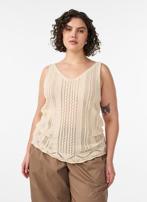 Frau mit lockigem Haar, die ein ärmelloses beiges Stricktop und weite braune Hosen trägt, zeigt ein Katzentattoo auf ihrem linken Arm.