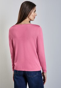 Růžový svetr s dlouhými rukávy, kulatým výstřihem a žebrovaným lemem. Nosené s tmavě modrými džíny, látka vypadá měkce s hladkou strukturou.