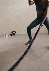 Schwarzes Battle Rope, gehalten von einer Person in dunkelgrüner Sportbekleidung. Der Boden ist hellgrau; kleine Hanteln befinden sich im Hintergrund und schaffen eine Fitnessstudio-Atmosphäre.
