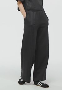 Osoba nosící volné černé kalhoty a tenisky Adidas s bílými pruhy, stojící s rukama v kapsách před jednobarevným pozadím.