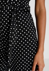 Svart jumpsuit med vita prickar, med en knytning i midjan, slät textur och figurnära silhuett.