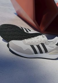 Baskets Adidas blanches et grises avec des rayures noires, placées sur une surface grise près d'une grande structure géométrique rouge.