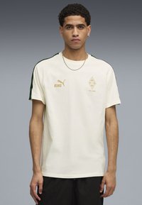 Ung mand iført en creme Puma t-shirt med "KING" og Borussia Mönchengladbachs "125 Jahre" logoer, stående foran en ensfarvet grå baggrund.