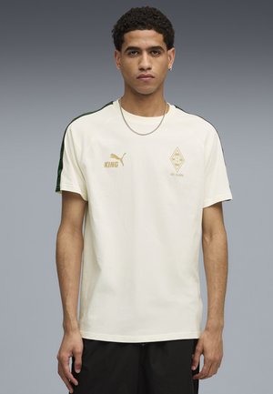 Ung mand iført en creme Puma t-shirt med "KING" og Borussia Mönchengladbachs "125 Jahre" logoer, stående foran en ensfarvet grå baggrund.