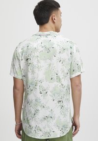 Camisa de manga corta en blanco con patrón floral verde; tejido ligero, corte relajado, detalle en el yugo trasero y dobladillo redondeado.