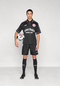 Μαύρη ποδοσφαιρική εμφάνιση με πουκάμισο με γιακά και κίτρινες λεπτομέρειες, λευκή σήμανση, συνδυασμένη με σορτς, κάλτσες και μαύρα παπούτσια ποδοσφαίρου. Κρατάει μια μπάλα με σχέδιο.