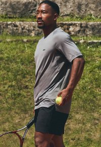 Camiseta atlética gris de manga corta con un logo, combinada con shorts negros. Sosteniendo una pelota de tenis amarilla y una raqueta de tenis roja.