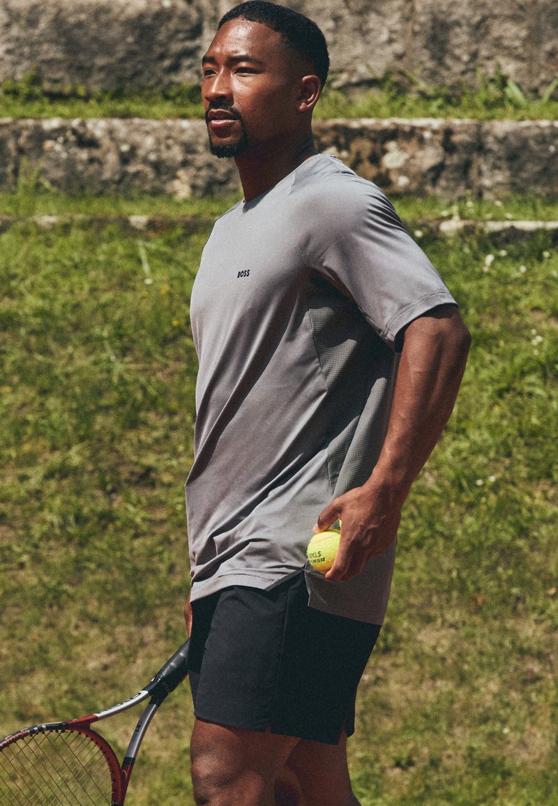 Camiseta atlética gris de manga corta con un logo, combinada con shorts negros. Sosteniendo una pelota de tenis amarilla y una raqueta de tenis roja.