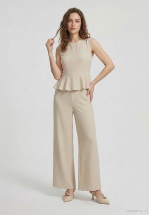 Donna con capelli mossi che indossa un top beige senza maniche con baschina, pantaloni beige a gamba larga coordinati e tacchi beige, in piedi davanti a uno sfondo semplice.