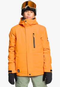 Veste isolée orange avec une poche zippée, une capuche réglable et des accessoires en noir. Portée avec des gants noirs et un bonnet.