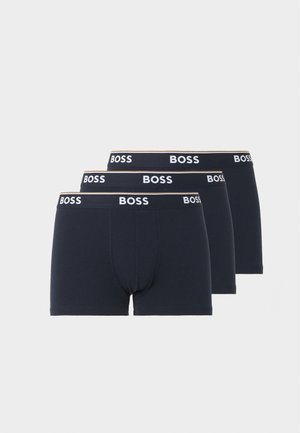 Muskulárny muž v tmavomodrých boxer nohavičkách s pásom "BOSS", stojaci na boso na bielom jednofarebnom pozadí, hľadiaci do strany.