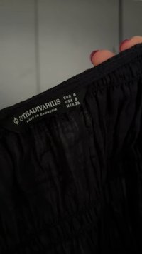 Črna obleka z blagovno znamko Stradivarius, ki kaže velikost EUR 8, USA 6, MEX 26, ki jo drži roka z našprejanimi nohti proti zamegljenemu ozadju.