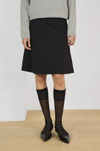 Jupe noire en ligne A avec une texture lisse, associée à des chaussettes noires à motifs arrivant jusqu'aux genoux et des chaussures noires à talons pointus. Pull gris au-dessus.