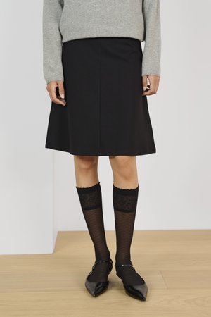 Jupe noire en ligne A avec une texture lisse, associée à des chaussettes noires à motifs arrivant jusqu'aux genoux et des chaussures noires à talons pointus. Pull gris au-dessus.