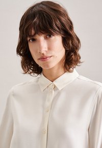 Cremfarbene Bluse mit kragenbehandeltem Hals, Knopfleiste und glatter Textur. Sie hat lange Ärmel und dezente Steppnähte.