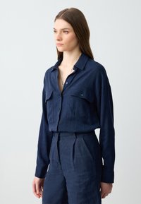 Marineblaue Leinenbluse mit Knopfleiste, zwei Brusttaschen und langen Ärmeln, kombiniert mit passenden hoch taillierten Hosen.