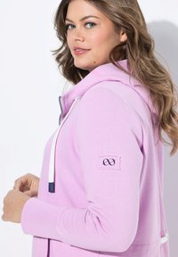 Haut à capuche violet clair avec une fermeture éclair à l'avant, des accents de cordon et un patch logo brodé sur la manche. Texture douce et design décontracté.
