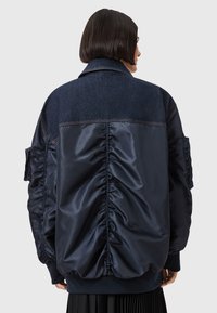Donna dai capelli corti e scuri indossa una giacca oversize blu navy con tessuto arricciato e yoke in denim, in piedi contro uno sfondo neutro.