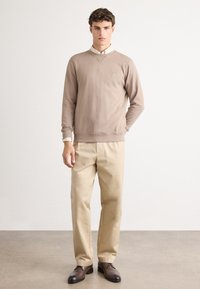 Sweatshirt marron clair avec un col rond et une patte de logo, associé à un pantalon beige et des chaussures marron foncé. Texture lisse, coupe décontractée.