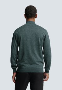 Homme vu de dos portant un pull texturé vert foncé et un pantalon noir sur un fond uni gris clair.