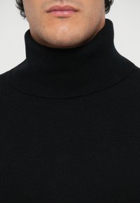 Schwarzer Rollkragenpullover, gerippte Textur, weicher Stoff, hoher Kragen. Nahaufnahme mit Fokus auf dem Halsbereich und den Details des Kleidungsstücks.