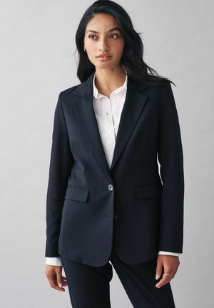 Navy blazer s klopami, se dvěma knoflíky a bočními kapsami. Nositý přes bílou košili s knoflíky, ukazující hladkou texturu látky.