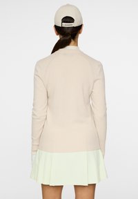 Maglione in maglia beige chiaro con orlo a costine e maniche lunghe, indossato su una gonna plissettata verde menta. Si vede un berretto beige da dietro.