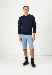 Marineblauwe sweatshirt met ronde hals, gedragen met lichtblauwe denim shorts, afgemaakt met zwarte sportschoenen en donkere sokken.