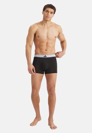 Uomo senza maglietta che sistema boxer neri con fascia in vita bianca e logo nero, in piedi a piedi nudi su uno sfondo chiaro uniforme.