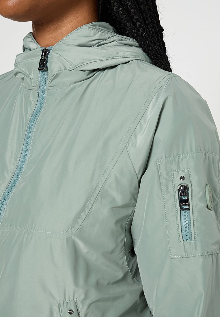 Hellgrüne Windjacke mit einer kapuzenformenden Reißverschluss, kontrastierendem blauen Reißverschluss und einer Reißverschlusstasche am linken Ärmel. Glatte Textur.