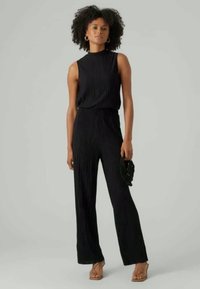 Melnā, bez piedurknēm kombinezonā ar augstu apkakli, ribotu struktūru, platām kājām un gludu audumu. Apvienots ar mazu, teksturētu kluču.