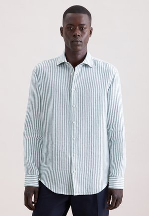 Mężczyzna w białej i jasnozielonej koszuli w pionowe paski na guziki oraz ciemnych spodniach, stoi na tle jednolitego jasnego tła.