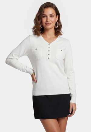 Vrouw die een witte, lange mouwen, geknoopte top met twee voorzakken en een zwarte rok draagt, staand met één hand op haar heup.