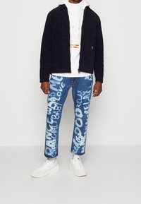 Navyblå kavaj med dragkedja, vit huvtröja med orange detaljer och blå jeans med vita textmönster. Matchas med vita sneakers.