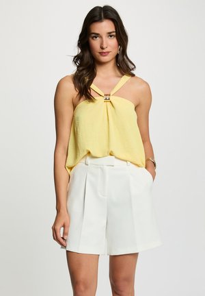 Top giallo senza maniche con centro arricciato e decorazione dorata, abbinato a pantaloncini bianchi su misura con pieghe e tasche laterali.