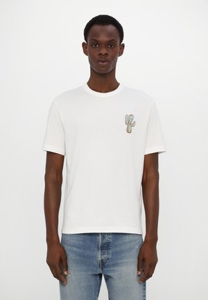 T-shirt bianco in cotone con collo a giro, caratterizzato da un ricamo colorato di un cactus sul lato sinistro del petto. Indossato con jeans azzurri chiari strappati.