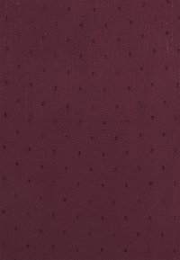 Bordeauxfarbener Stoff mit glatter Textur, der kleine, erhaben genoppte Punkte aufweist, die gleichmäßig verteilt sind. Das Material wirkt leicht und vielseitig.